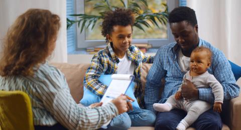 A black father sits on a couch with his toddler on his lap and his young son next to him while they talk to a woman who's listening and writing on a clipboard