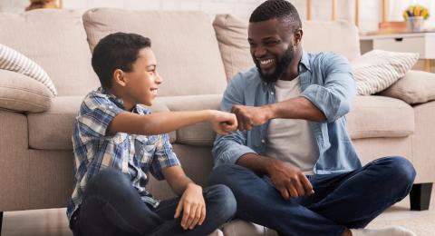 a dark skinned man and a medium light skinned young teen boy fist bump while sitting on the floor in front of a couch