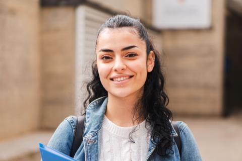 a young woman or youth smiles for the camera, she has medium-light skin, long curly black hair and is wearing a backpack and holding a binder.
