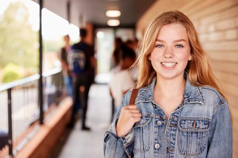 A young blond female-presenting teenager poses for a photo in what looks like a highschool balcony. Other students with backpacks can be seen talking together in the background.