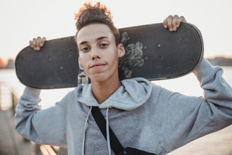 a male-presenting young person with medium-light skin and short styled hair in a hoodie holds a skateboard on their shoulder behind them